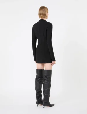 Back view of a person wearing a black long-sleeve mini dress and over-the-knee black leather boots with stiletto heels on a white backdrop.