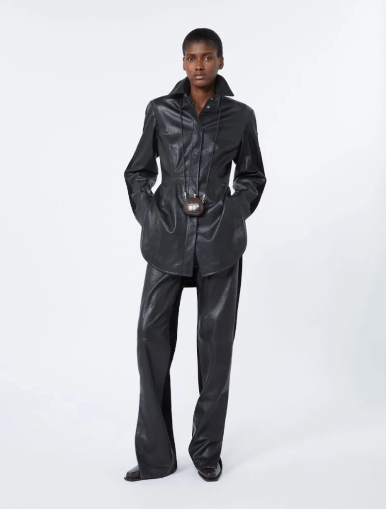 Model in a black leather-look outfit: long shirt with wide-leg pants and a small brown belt bag at the waist, studio background.