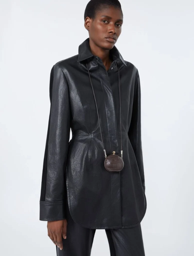 Model wearing a dark, shiny leather-look coat with a small brown mini bag hanging from a strap around the neck.