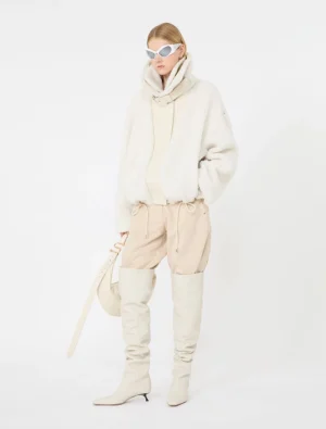 Model wearing an off-white fuzzy coat, beige jogger pants, and over-the-knee boots with white sunglasses and a light strap bag.