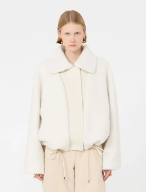 Model wearing a white shearling jacket with a wide collar over a cream blouse and beige drawstring trousers, facing the camera.