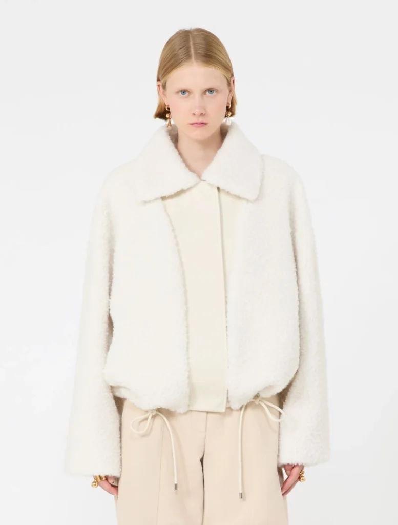 Model wearing a white shearling jacket with a wide collar over a cream blouse and beige drawstring trousers, facing the camera.
