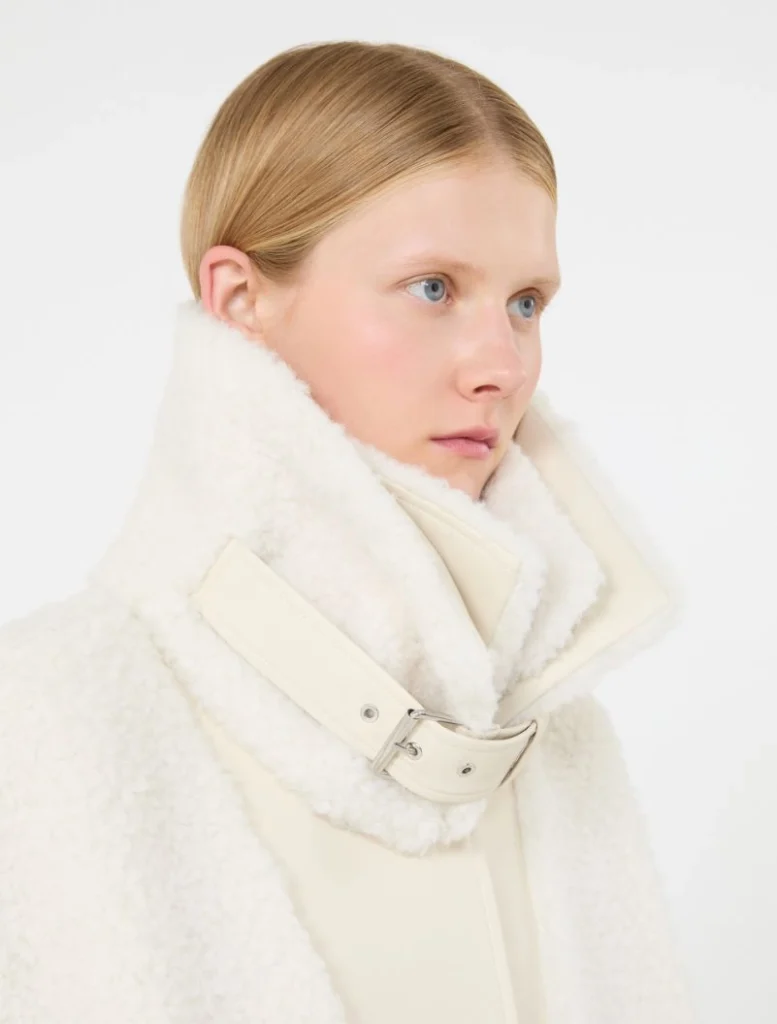 Young woman with blonde hair wearing a white shearling coat with a large high collar.