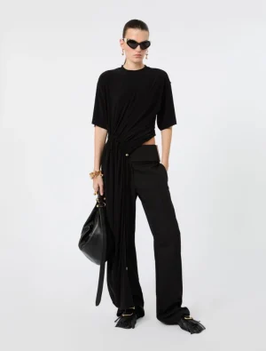 Model in an all-black ensemble: twisted draped top with a long sash, wide-leg pants, black handbag, and gold jewelry with sunglasses.