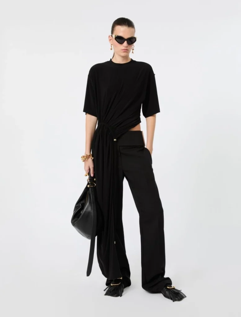 Model in an all-black ensemble: twisted draped top with a long sash, wide-leg pants, black handbag, and gold jewelry with sunglasses.