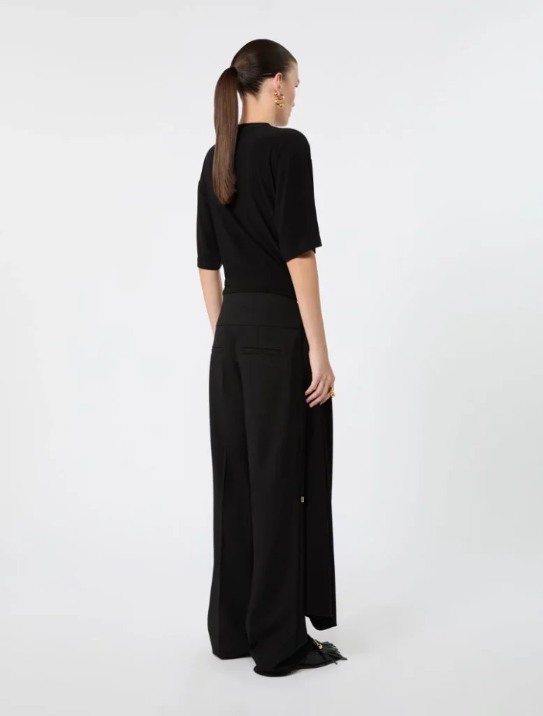 Woman seen from behind in an all-black outfit: a loose black top and wide-leg black trousers with back pockets, wearing gold earrings and a ponytailed hairstyle.
