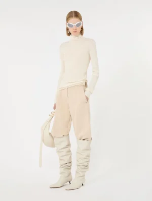 Female model in a cream turtleneck and beige slouchy pants with tall cream boots, white cat-eye sunglasses, and a light bag against a white studio background.