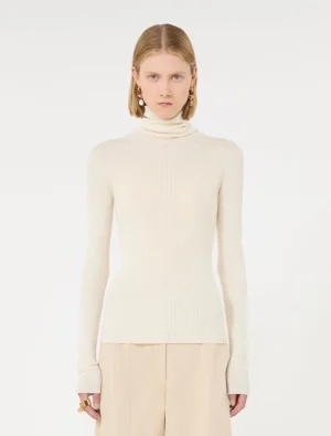 Model wearing a cream ribbed turtleneck sweater with beige wide-leg pants, accessorized with gold earrings and rings against a white background.