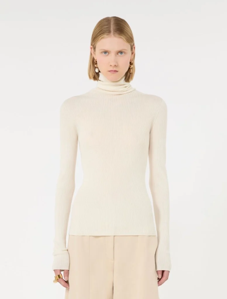 Model wearing a cream ribbed turtleneck sweater with beige wide-leg pants, accessorized with gold earrings and rings against a white background.
