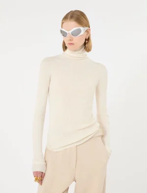 Model in a cream ribbed turtleneck sweater and beige trousers, wearing white angular sunglasses against a white backdrop.