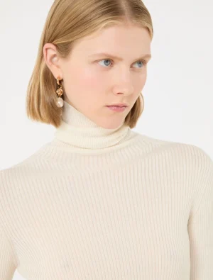 Portrait of a woman with short blonde hair wearing a cream ribbed turtleneck and gold earrings, looking to the right far.