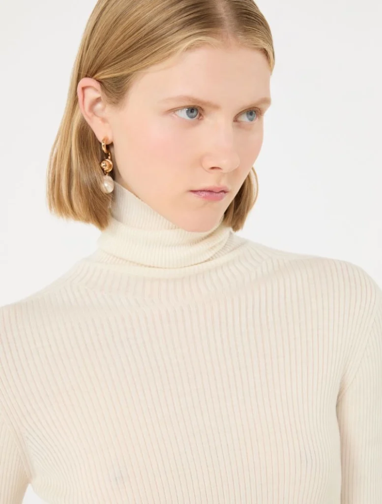 Portrait of a woman with short blonde hair wearing a cream ribbed turtleneck and gold earrings, looking to the right far.