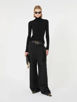 Model in an all-black outfit: black turtleneck, wide-leg trousers, gold belt, black clutch, sunglasses.