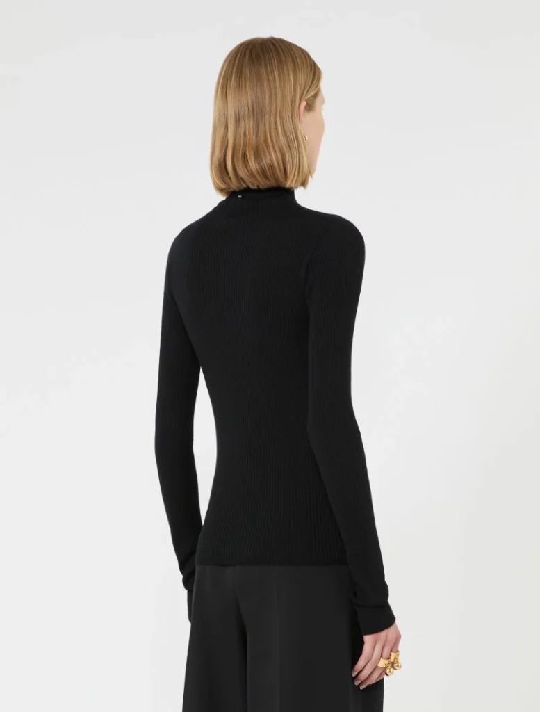Back view of a person wearing a black ribbed turtleneck sweater and black wide-leg pants, blonde hair cut to the shoulders.