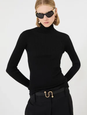 Model in a black ribbed turtleneck and black trousers with a gold-buckle belt, wearing dark sunglasses and pearl earrings.