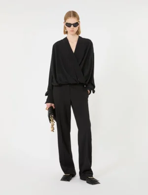 Model wearing an all-black look: wrap-style draped top with wide-leg pants, black sunglasses, and a black clutch with a chunky gold chain, standing against a white backdrop.