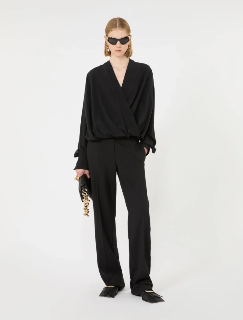 Model wearing an all-black look: wrap-style draped top with wide-leg pants, black sunglasses, and a black clutch with a chunky gold chain, standing against a white backdrop.