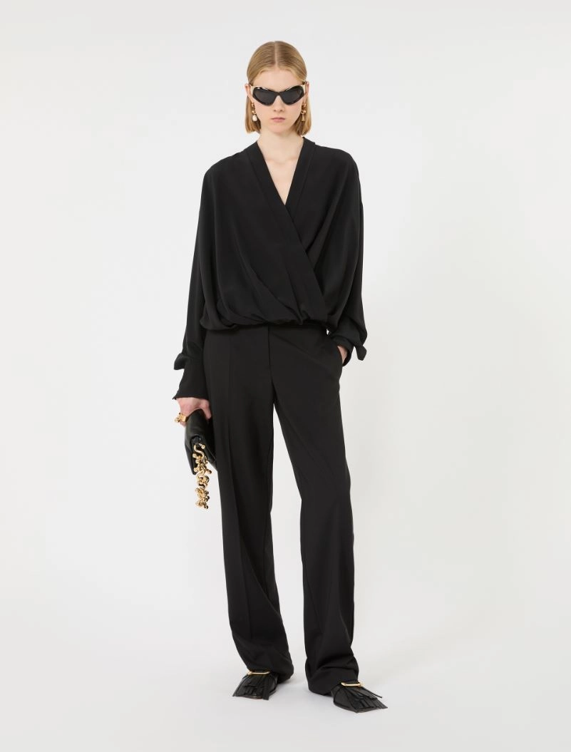 IMG_3802 - New Trend Model wearing an all-black look: wrap-style draped top with wide-leg pants, black sunglasses, and a black clutch with a chunky gold chain, standing against a white backdrop.