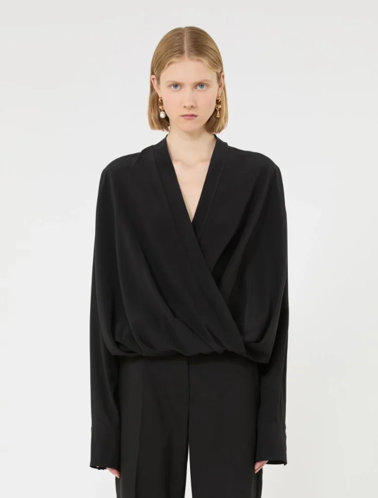 Young woman wearing a black draped wrap blouse with wide sleeves and matching black pants, neutral studio backdrop