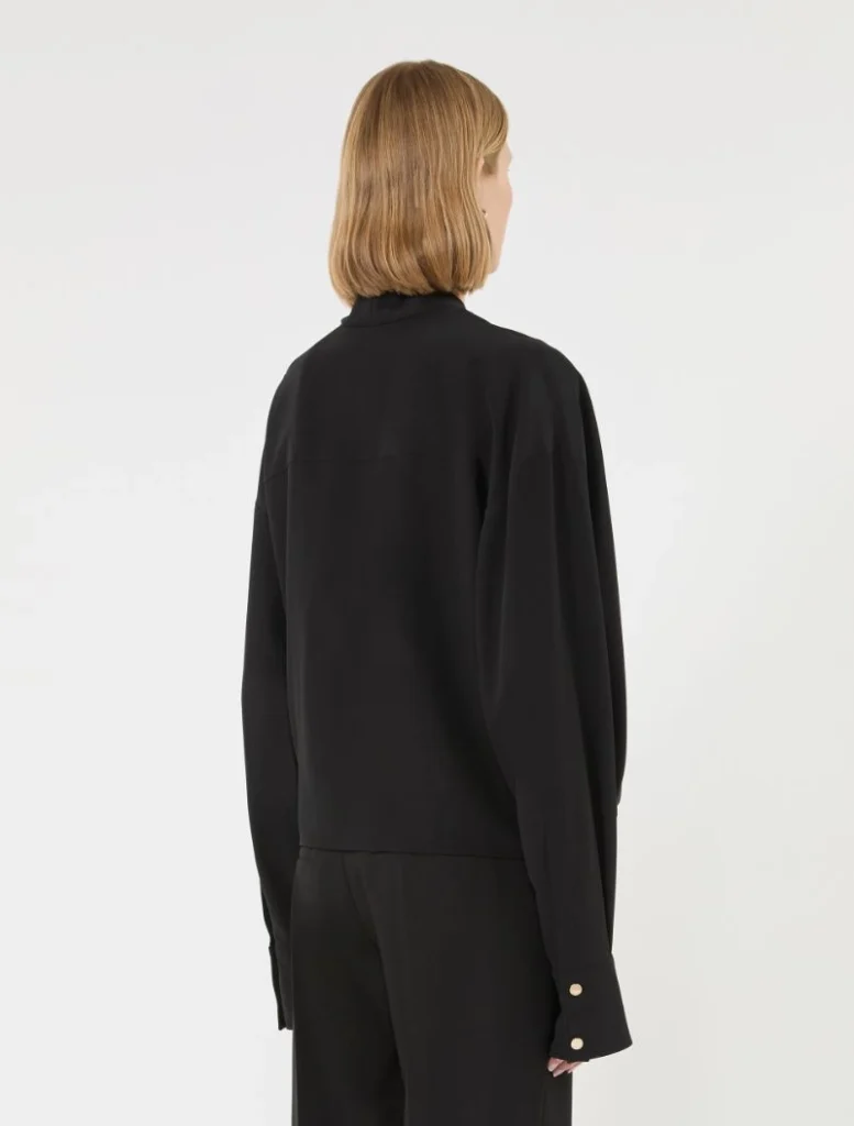 Back view of a person wearing a black long-sleeve jacket with gold cuff buttons, blonde bob haircut, neutral studio background.