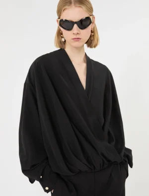 Model wearing a black, draped wrap blouse with oversized sleeves and matching pants, accessorized with large sunglasses and gold earrings.