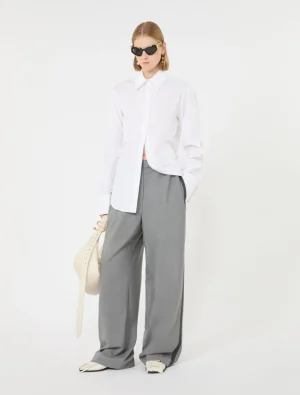 Model wearing an oversized white button-down shirt with gray wide-leg trousers, holding a cream hat and wearing black sunglasses.