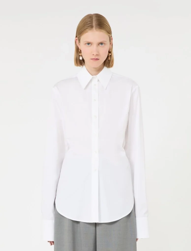 Model wearing a white button-down shirt with long sleeves and a curved hem, paired with gray trousers, facing the camera.