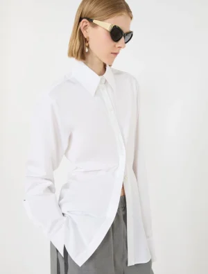 Model wearing an oversized white button-down shirt with gray trousers, sunglasses, and gold earrings in a minimalist studio.