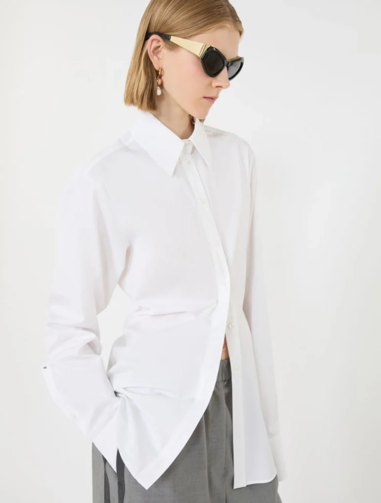 Model wearing an oversized white button-down shirt with gray trousers, sunglasses, and gold earrings in a minimalist studio.