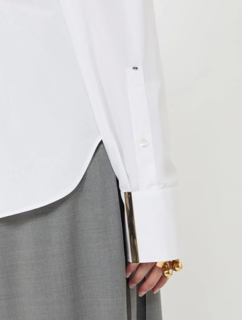 Close-up of a white dress shirt cuff with a gold metal cuff bracelet and gold rings, worn over a gray skirt.