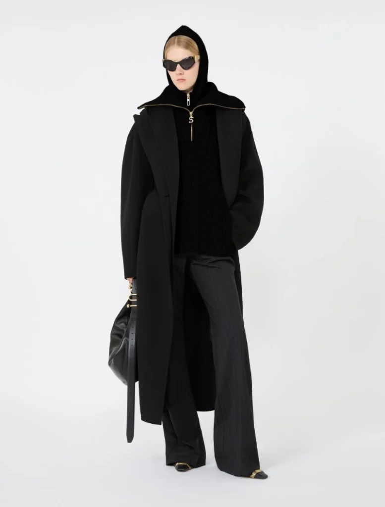 Model in an all-black, oversized coat with hood, sunglasses, and a black handbag for a high-fashion look.