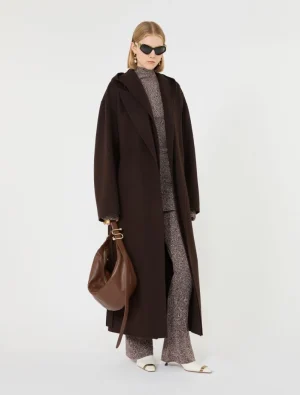 Model wearing a long dark brown coat over a textured knit set, with white sandals and a brown bag, plus black sunglasses.