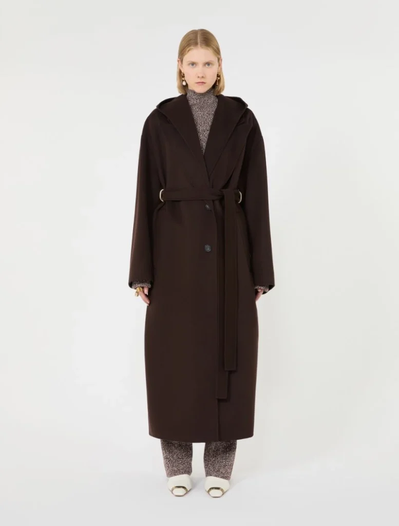 Full-length view of a model wearing a dark brown, belted wool coat with wide lapels, paired with speckled pants and white shoes