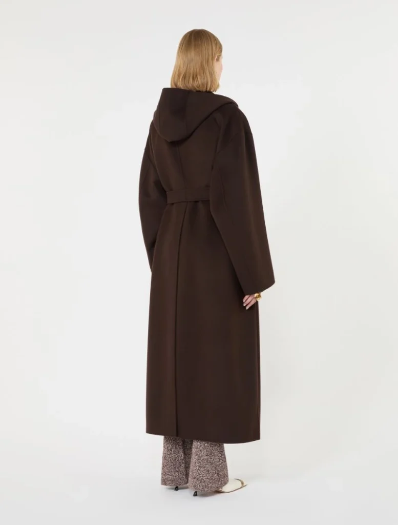 Back view of a person wearing a long brown coat with a belt and hood, standing against a plain white backdrop.