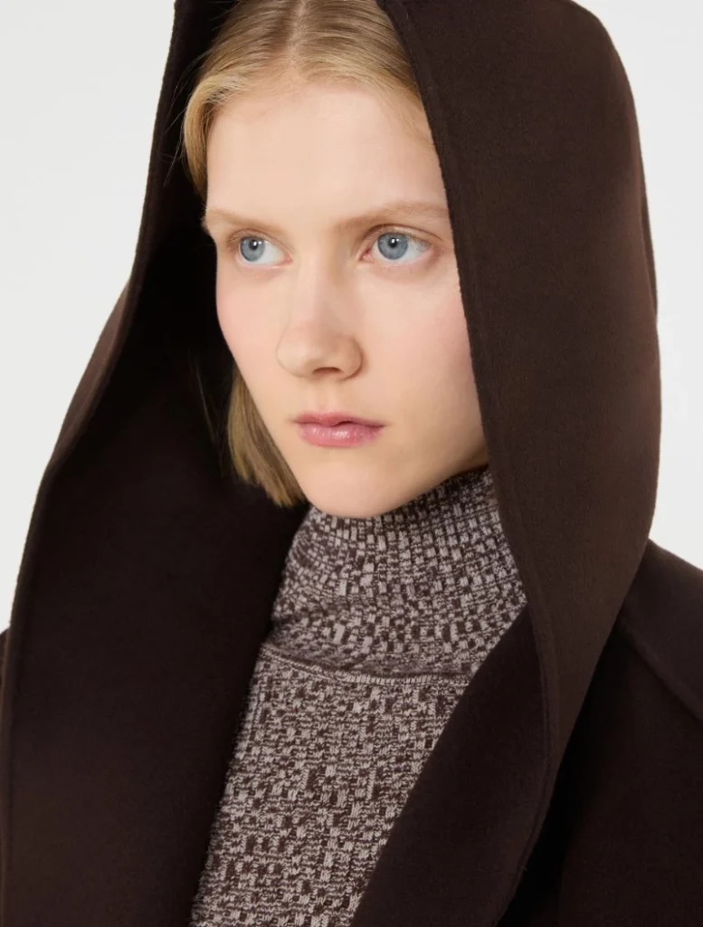 Young woman with blue eyes wearing a dark brown hooded coat and a patterned turtleneck, looking to the side.
