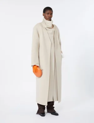 Model wearing a long beige coat over a cream turtleneck, with bright orange gloves and matching orange clutch, brown trousers and black shoes on a white backdrop.
