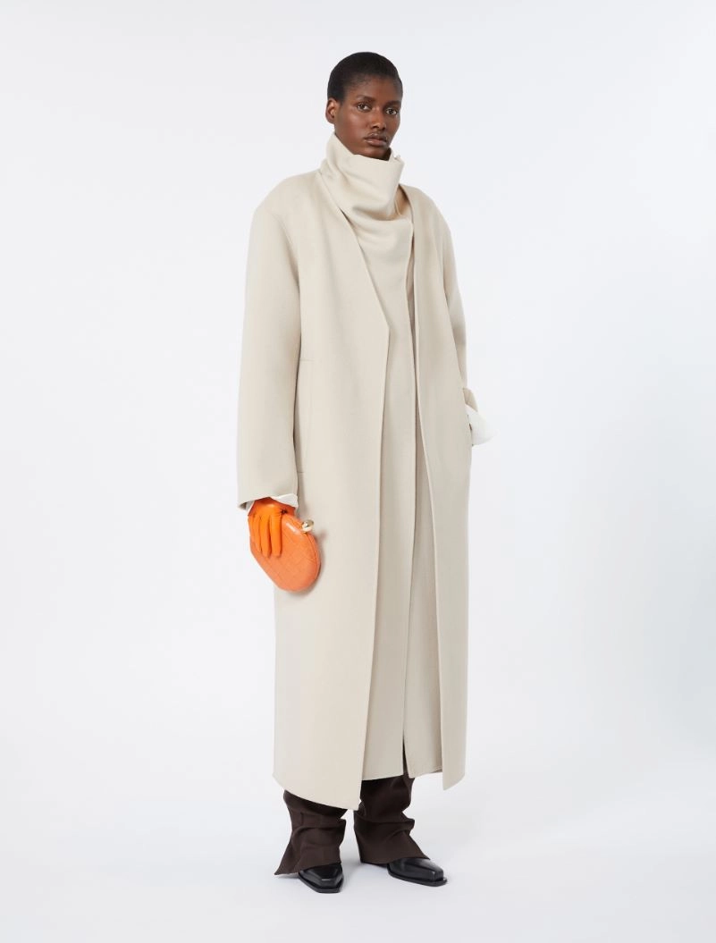 IMG_3825 - New Trend Model wearing a long beige coat over a cream turtleneck, with bright orange gloves and matching orange clutch, brown trousers and black shoes on a white backdrop.