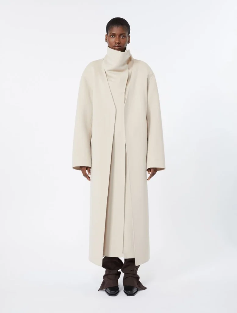 Full-body shot of a model wearing a long, cream-colored oversized coat with a high collar, standing against a white background.