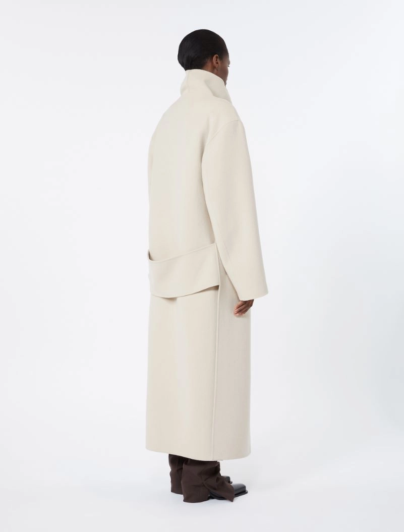 IMG_3827 - New Trend Back view of a person wearing a long cream coat with a high collar and a waist belt, paired with brown boots.
