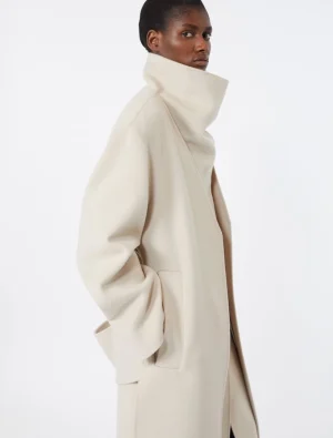 Model wearing a cream oversized wool coat with a tall stand collar, shown from the side with hands in pockets.