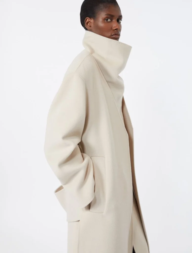 Model wearing a cream oversized wool coat with a tall stand collar, shown from the side with hands in pockets.