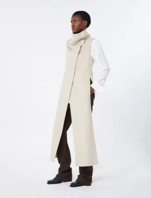 Model wearing a long beige sleeveless coat over a white shirt and brown wide-leg pants.