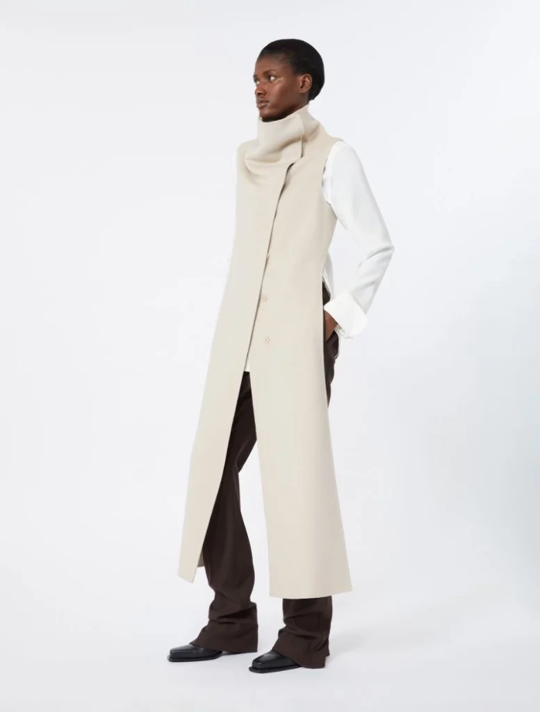 Model wearing a long beige sleeveless coat over a white shirt and brown wide-leg pants.