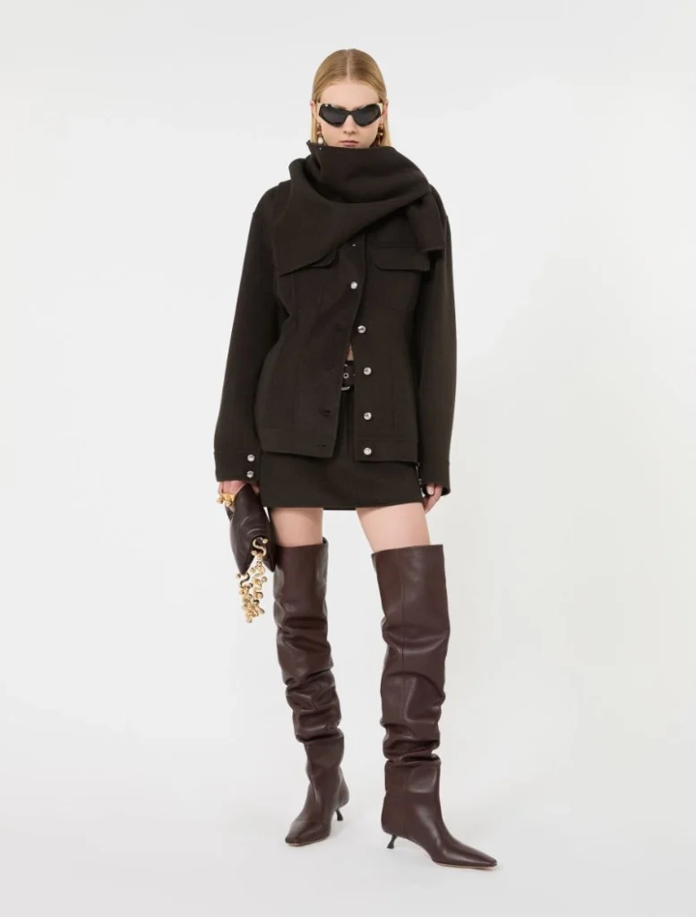 Model wearing an oversized black coat with a large scarf, black mini skirt, tall brown knee-high boots, sunglasses, and a black clutch with a gold chain.