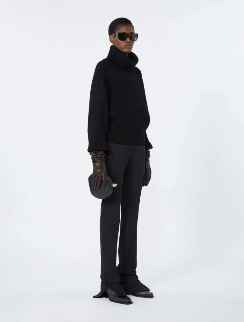 Model in an all-black ensemble: oversized turtleneck, wide-leg pants, brown gloves, sunglasses, and a small black handbag used as a fashion look.