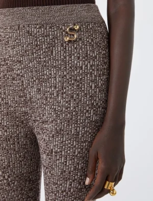 Close-up of brown tweed trousers with a gold S-shaped pin at the hip and a chunky gold ring on the wearer's finger.
