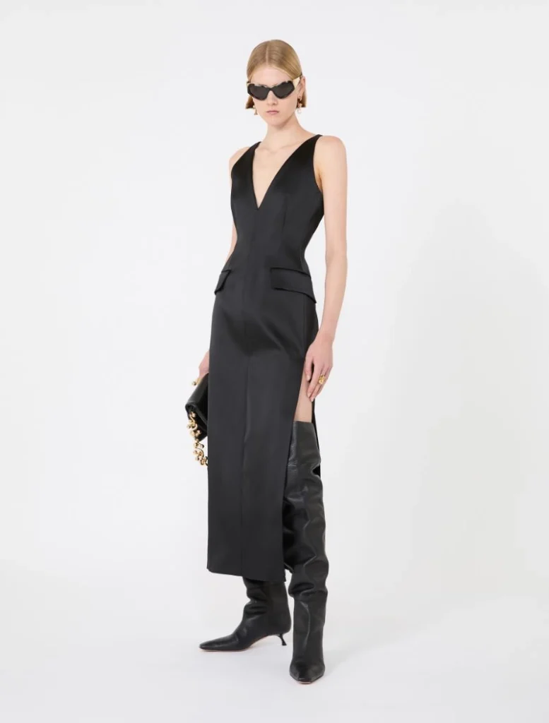 Model wearing a black sleeveless V-neck midi dress with a front slit, paired with knee-high black boots and sunglasses in a studio setting.