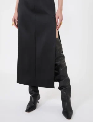 Lower half of a person wearing a black midi dress with a side slit and tall black boots.