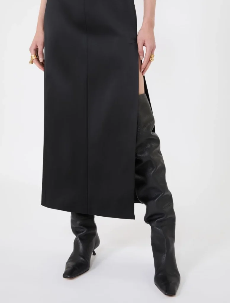 Lower half of a person wearing a black midi dress with a side slit and tall black boots.