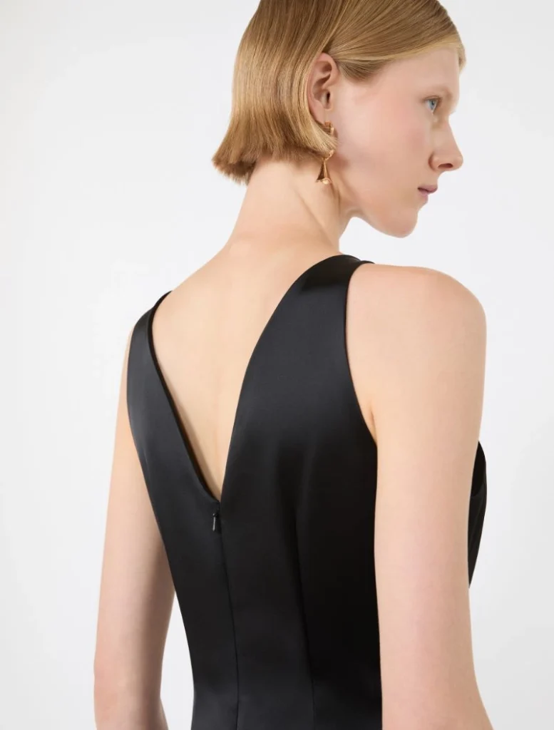 Back view of a woman in a black sleeveless dress with a V-shaped back and a center zipper; short blonde hair and gold earrings, neutral studio background.